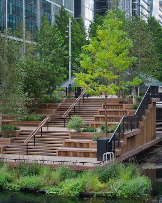 A modern urban garden scene in Canary Wharf featuring a small grassy lawn bordered by paved pathways, with several young trees and trimmed hedges surrounding the area. The garden includes lampposts along the pathways and a few benches where people are seated, enjoying the outdoor space. In the background, tall contemporary skyscrapers with reflective glass facades tower over the garden, showcasing a mix of geometric and curvilinear architectural designs. The sky overhead is partly cloudy with patches of blue, and natural light illuminates the greenery and building exteriors, highlighting the textures of wet soil, neatly mowed grass, and well-maintained plant borders. Gardeners canarywharf.org.uk offers insights into maintaining such landscaped areas, emphasizing lawn care, hedge trimming, and general garden maintenance amidst the urban environment.