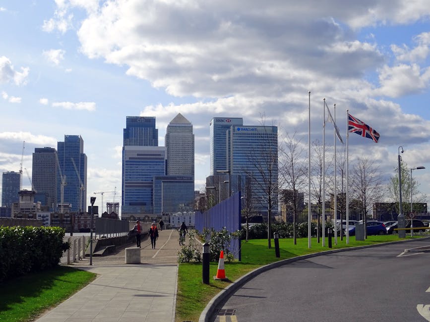 A landscaped urban promenade with a paved walkway lined by neatly trimmed grass and small shrubs leading towards a backdrop of modern high-rise office buildings in Canary Wharf. Several flagpoles with Union Jack flags are visible, along with leafless young trees along the edge of the path. A few pedestrians are strolling along the pathway on a partly cloudy day, with the city’s skyscrapers, including those with HSBC and Barclays signage, prominently visible under a partly cloudy sky, illustrating a blend of outdoor garden maintenance and cityscape landscaping. Gardeners Canary Wharf provides professional outdoor care and landscaping work in this area, ensuring a well-maintained, visually appealing environment amidst the urban setting.