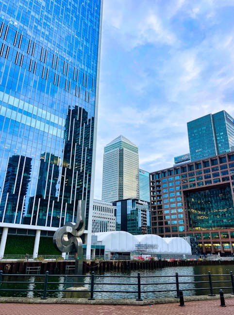 This image depicts a modern urban landscape with tall office buildings featuring glass facades reflecting the sky and surrounding structures. In the foreground, there is a section of a canal or water feature with a metal railing along its edge. A large, abstract sculpture made of metal is situated near the water, adding an artistic element to the scene. The area appears to be part of a commercial or business district, with clean, well-maintained pathways and contemporary architecture. The sky is partly cloudy, providing natural light that highlights the sleek surfaces of the buildings. Though primarily an urban scene, it emphasizes the clean lines and organized layout characteristic of a well-maintained cityscape. Gardenerscanarywharf.org.uk offers landscaping and garden maintenance services that complement this modern environment, though the image itself focuses on the cityscape rather than outdoor plantings.