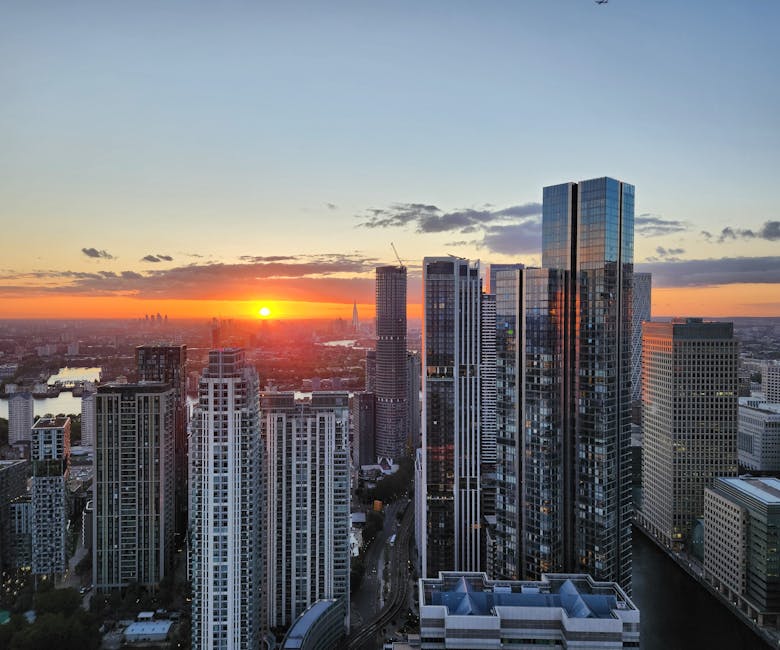 A panoramic view of a modern cityscape at sunset, showcasing numerous tall, glass-clad skyscrapers with reflective surfaces capturing the warm hues of the setting sun. The foreground features a cluster of high-rise buildings with sleek, contemporary designs, some with visible rooftop structures. In the background, a river winds through the city, bordered by more buildings and trees, with the horizon glowing in vibrant orange and yellow tones. The sky is partly cloudy with soft, wispy clouds, and the natural light highlights the textures of the glass facades, creating a striking contrast between the urban environment and the warm, fading daylight. This scene reflects an urban landscape that would be typical of a thriving financial or business district, with the overall atmosphere calm and picturesque as evening approaches.