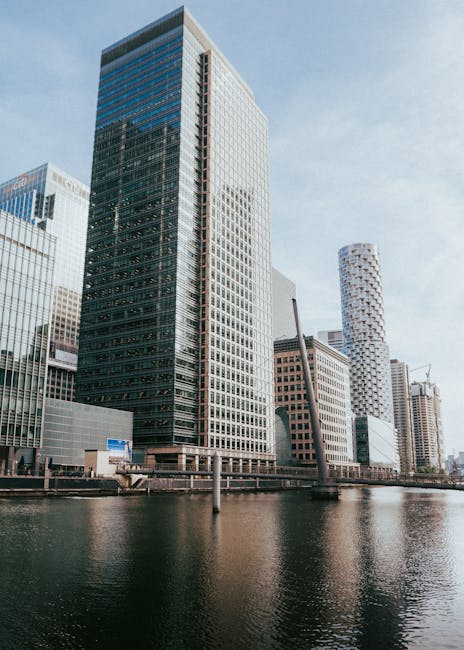 A panoramic view of a modern cityscape at sunset, showcasing numerous tall, glass-clad skyscrapers with reflective surfaces capturing the warm hues of the setting sun. The foreground features a cluster of high-rise buildings with sleek, contemporary designs, some with visible rooftop structures. In the background, a river winds through the city, bordered by more buildings and trees, with the horizon glowing in vibrant orange and yellow tones. The sky is partly cloudy with soft, wispy clouds, and the natural light highlights the textures of the glass facades, creating a striking contrast between the urban environment and the warm, fading daylight. This scene reflects an urban landscape that would be typical of a thriving financial or business district, with the overall atmosphere calm and picturesque as evening approaches.